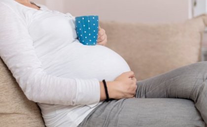 A woman in a white top reclines on a couch, resting a blue coffee cup on her pregnant stomach